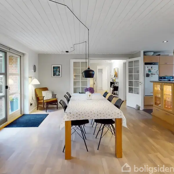 Langbord med seks stole står i spisestue med lyst trægulv og hvide vægge. En lampe hænger over bordet, og en dør står åben til køkkenet. Tekst: "RASM".