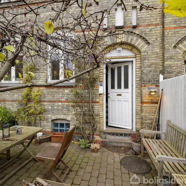 Et gammelt murstensrækkehus med en hvid dør. Foran er der et bord med stole under et træ. Små planter omkranser området. Addressen er "22".