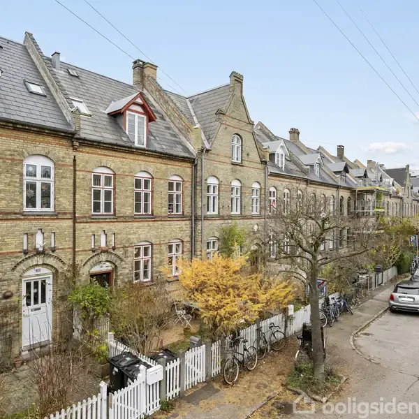 En række ældre byhuse i mursten står langs en stille vej med parkerede biler. Haverne er adskilt af hvide stakitter, og cykler er placeret ved fortovet.