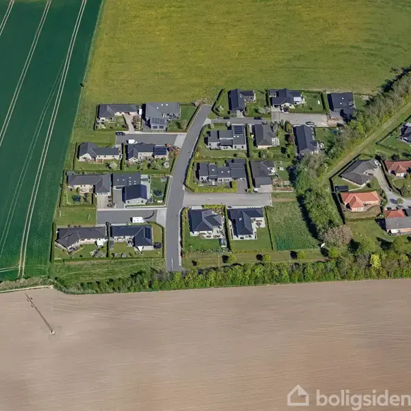Boligområde viser huse med mørke tage arrangeret i et mønster af veje. Omgivet af grønne marker og træer, ligger hytterne i et landligt miljø med åbne marker omkring.