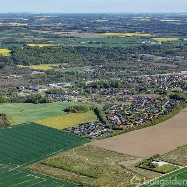 Landskab viser en by med huse og veje omgivet af store marker og skove. Store, åbne marker strækker sig til horisonten, med spredte skovområder og landbrugsmarker i forskellige farver.