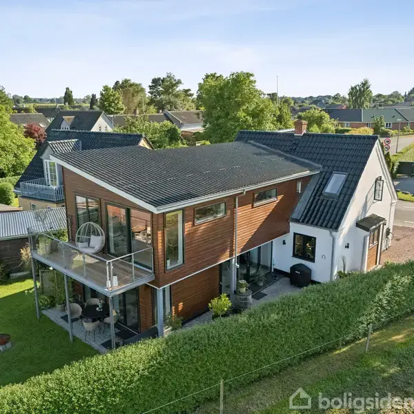 Moderne hus med glasfacade og balkon. Huset står i en velplejet have omkranset af grønne hække. Flere træer og nabohuse omgiver området, der er roligt og grønt.