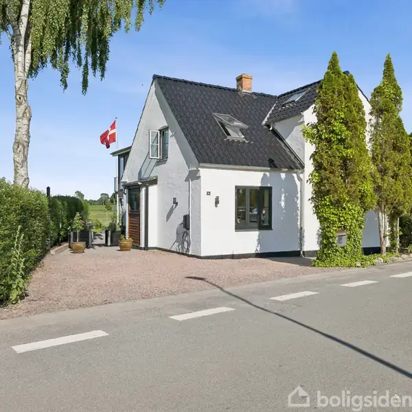 Hvidt hus med sort tag står ved en vej, omgivet af træer og hække. En dansk flagstang vifter i vinden i haven ved siden af huset.