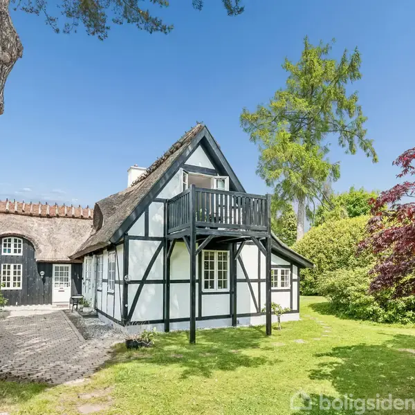 Bindingsværkhus med stråtag og balkon står i en frodig have med grønne træer og buske omkring. Sollys belyser facaden, mens en stensti leder mod indgangen.