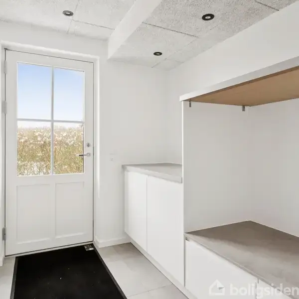 En hvid entré med en dør til venstre fører ud til haven. Til højre er der et åbent skab med hylder og kroge. Værelset er lyst og moderne.