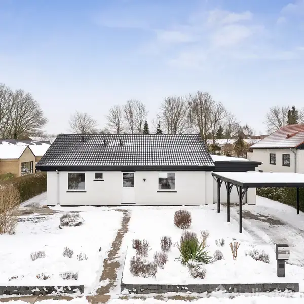 Hvidt hus står stille i snedækket have med stier og carport. Omgivet af hække og naboens huse under en klar, blå himmel.