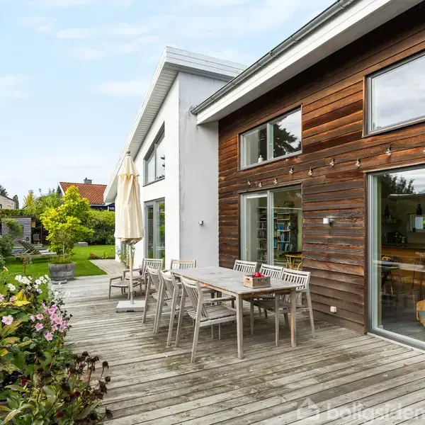 Et moderne hus med store vinduer har en træterrasse. Et træbord med stole står på terrassen, omgivet af grønne planter og blomster, med en parasol i baggrunden.