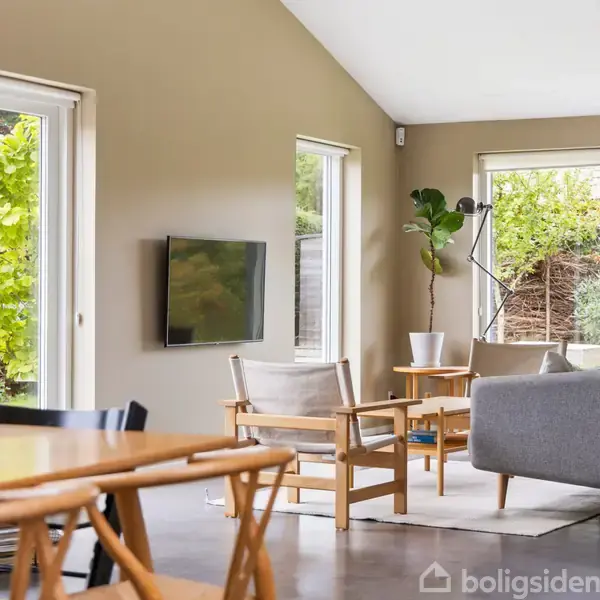 Stue med grå sofa og to stole, hvor et fjernsyn hænger på væggen. Store vinduer og glasdør åbner mod en grøn have, mens spisebordet står i forgrunden.