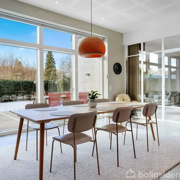 Et spisebord med seks stole er placeret på et tæppe i en lys spisestue. Store vinduer og en glaspaneldør viser en terrasse og have udenfor.