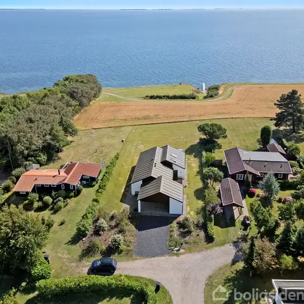 Et hus med gråt tag står foran en kystlinje ved havet, omgivet af grønne marker og mindre huse. Landskabet er frodigt med træer og buske omkring bygningerne.