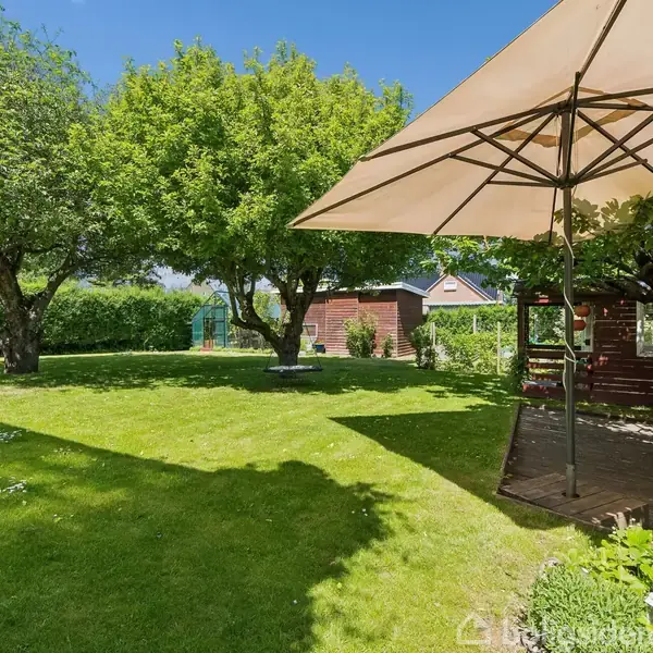 Parasol står åben i en grøn have med frodige træer. I baggrunden ses et rødt hus, en græsplæne og en hækkant. Solskin kaster skygger på plænen.