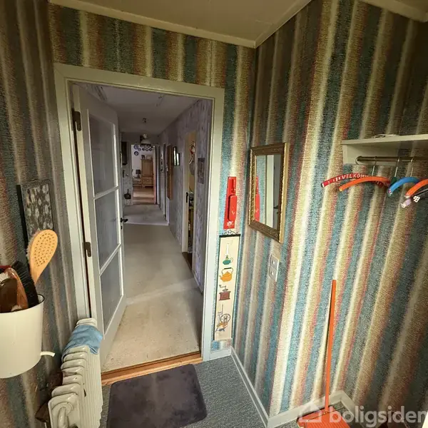 En snæver gang er dekoreret med stribede tapeter. Væghylder holder farverige bøjler. Et spejl hænger på væggen. En dør fører videre ind i huset.