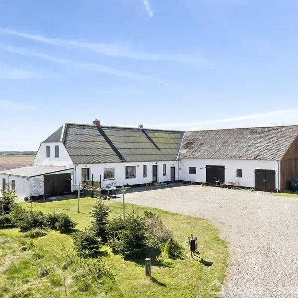 Et hvidt hus med gråt tag står stille på en grusvej omgivet af grønne marker og træer under en klar blå himmel.