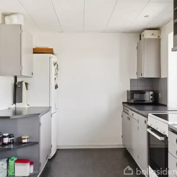 Et køkken med grå skabe og sort bordplade. Et køleskab står i hjørnet nær en håndvask. Krydderier er placeret på en hylde. En ovn er indbygget i højre side.