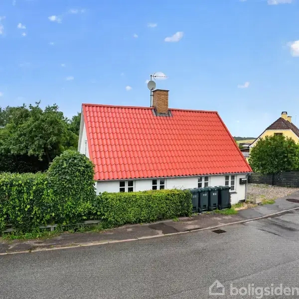 Et lille hvidt hus med rødt tag står ved en vej. Det er omgivet af grønne hække og buske under en blå himmel med enkelte skyer.
