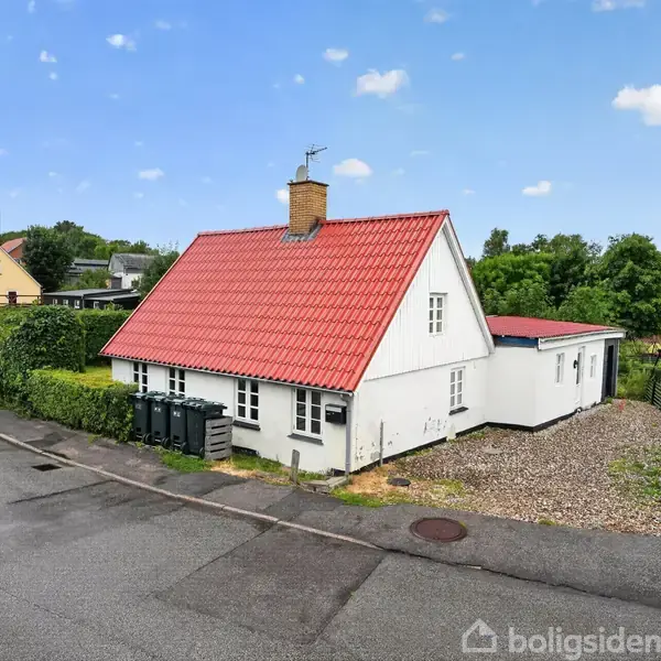 Huset har et rødt tegltag og hvid facade, står fredeligt på et hjørne med grønne buske omkring. Vejen forløber langs siden, og der er en grusplads ved siden.