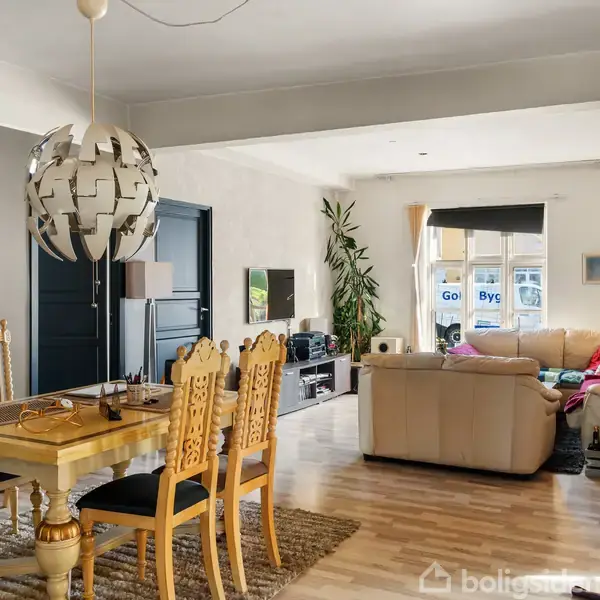 Et stilfuldt, åbent rum med spisebord og to stole, sofahjørne med beige sofa, farverige puder, plante og lamper. Store vinduer lyser rummet op. Ord på vinduer: "Go Byg".