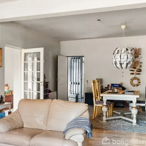 En beige sofa står i et stueområde med hvide vægge og trægulv. Et rundt spisebord med stole og dekorativ lampe hænger i midten. Åbne døre fører til andre rum.