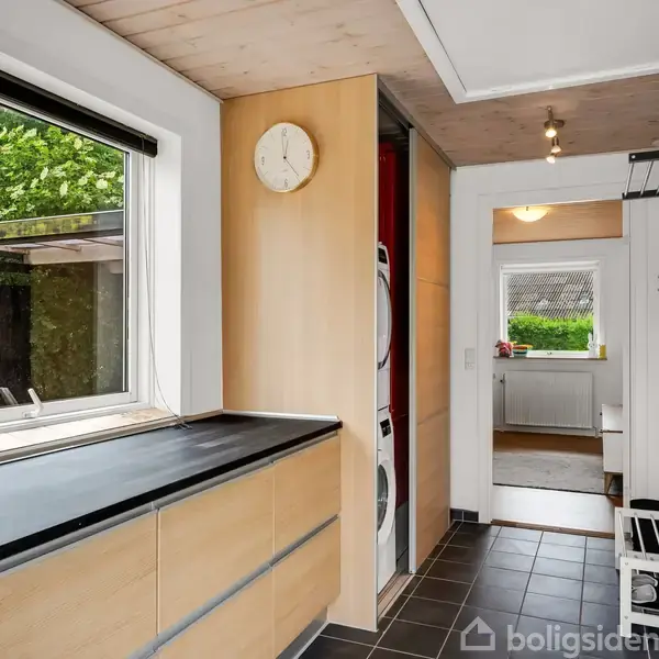 Et bryggersområde med sorte fliser har vaskemaskine og tørretumbler stablet i et skab. Et vindue til venstre viser ud til en have. En dør fører til et andet rum.