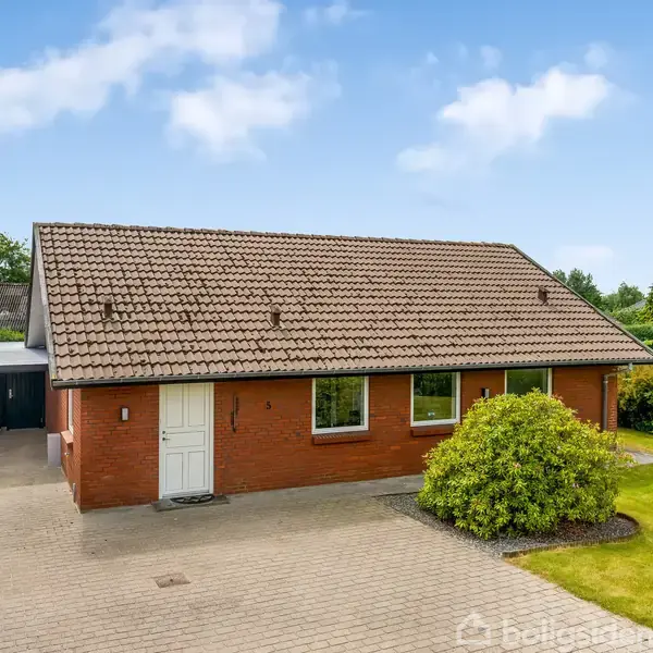 En rød murstensvilla med saddeltag, står stille omkranset af en velplejet have med græsplæne og buske, ved siden af en carport i et villakvarter.
