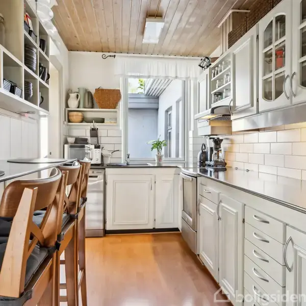 Et lyst køkken med hvide skabe og træbordplade. Barstole står ved en høj disk. Hylder med køkkenartikler hænger på væggen. Et vindue giver lys, med grønne planter udenfor.