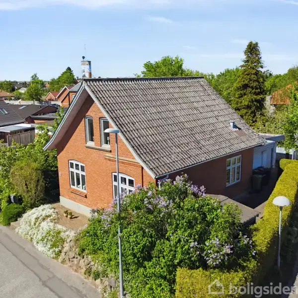 Et rødt murstenshus med skråtag står stille på en hjørnegrund omgivet af grøn beplantning i et beboelseskvarter med parkerede biler og andre huse i nærheden.