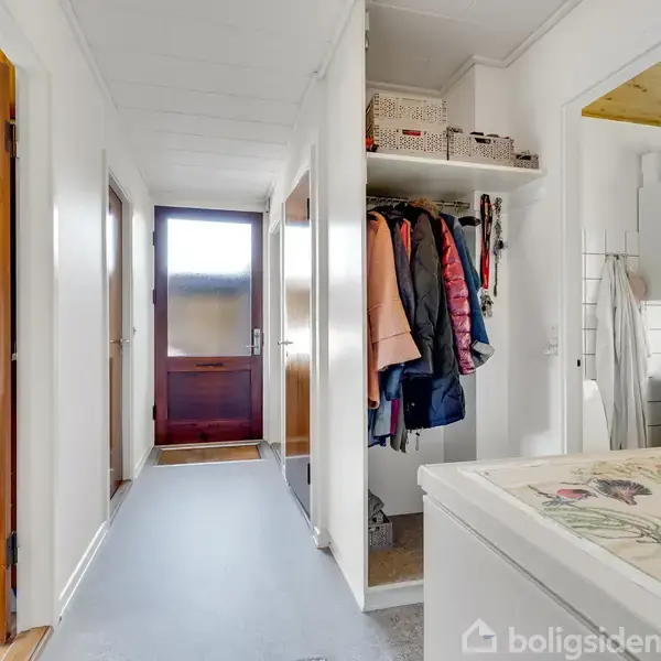 Gang med tøjskab, hvor jakker hænger. Skabet står mod en væg med åbning til et rum med vaskefaciliteter og skabe. Døre og lysindfald ses for enden af gangen.