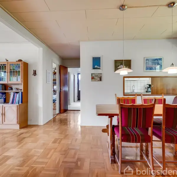 Spisebord med røde stole står i en stue med parketgulv. Bogreol til venstre, vindue med udsigt til have til højre. Malerier hænger på væggen.