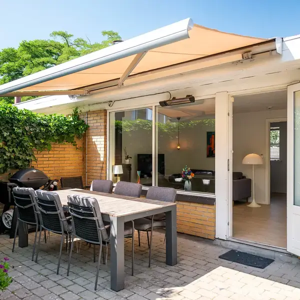 Terrasseområde med spisebord og stole, dækket af en markise. I baggrunden ses glasdøre, der åbner ind til en lys stue med sofa, lamper og dekorationer.