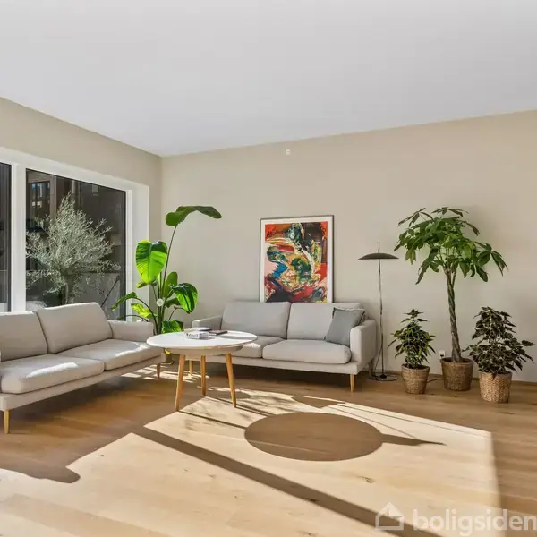 To sofaer står mod en lys væg med et farverigt maleri hængende over. Store vinduer til venstre lukker lys ind, og to grønne planter pynter rummet.