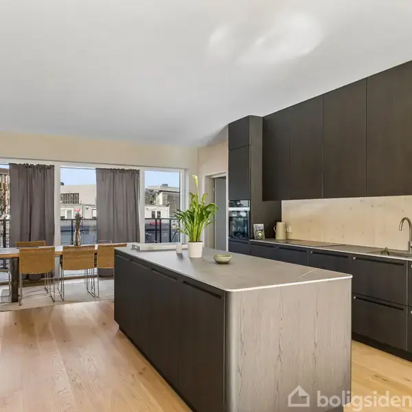 Et moderne køkken med en stor, mørk køkkenø, hvorpå der står en plante. Det er omgivet af grå skabe, og i baggrunden ses et spisebordsområde med udsigt til en balkon.