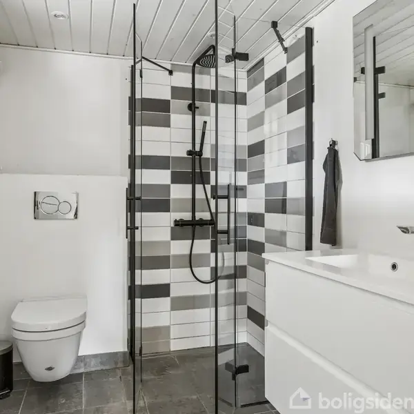 Badeværelse med bruser i glasafskærmning, moderne hvidt og sort flisebelagt vægdesign. Indeholder toilet, håndvask med skab, spejl og en lille skraldespand ved siden af toilettet.