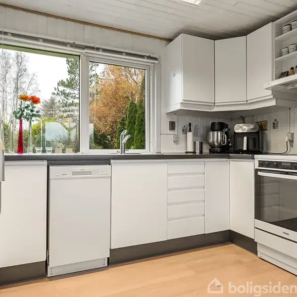 Hvidt køkken indeholder opvaskemaskine, kaffemaskine og ovn. Store vinduer viser efterårsfarver udenfor, og der står en vase med blomster i vindueskarmen. Trægulv og lyse skabe præger rummet.