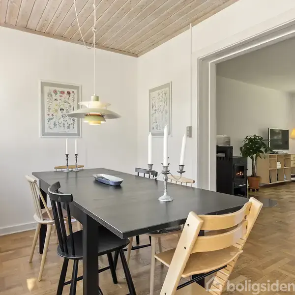Et spisebord med sorte og træstole omgivet af lyse vægge og trægulv. Bordet har lysestager, og der hænger en lampe over det. Væggene har indrammede billeder.