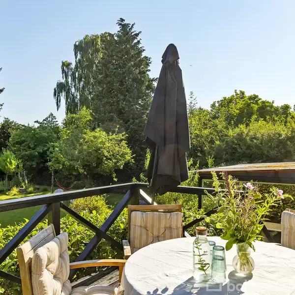 En rund bord med blomster og flasker står på en terrasse. Parasol står oprejst, sammenfoldet. Omgivet af behagelige stole, frodige træer og buske i en solrig have.