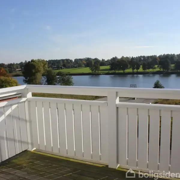 En hvid balkon med udsigt over en sø og grønne træer i baggrunden under en blå himmel.