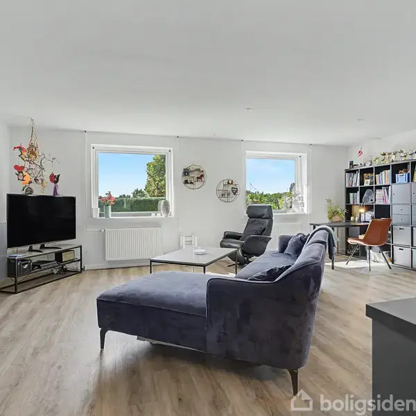 En stue har en blå sofa i centrum, med et tv og møbler på hver side. To vinduer giver lys, og der er flere billeder og reoler i rummet.