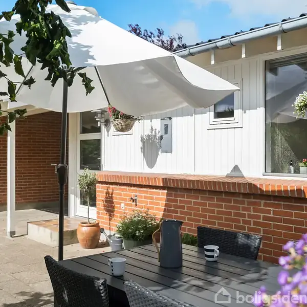 Parasol dækker bord og stole på terrasse. Hvide vægge og mursten bagved. Blomster i hængende krukker dekorerer omgivelserne. Klar himmel giver lys, hvilket skaber en indbydende atmosfære.