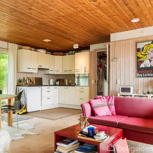 Sofa med lyserøde puder står i stue med åbent køkken. Rundt spisebord ved glasdør. Træmøbler og -loft. Kunstplakat med teksten "Louisiana Roy Lichtenstein" hænger på væggen. Bøger under bordet.