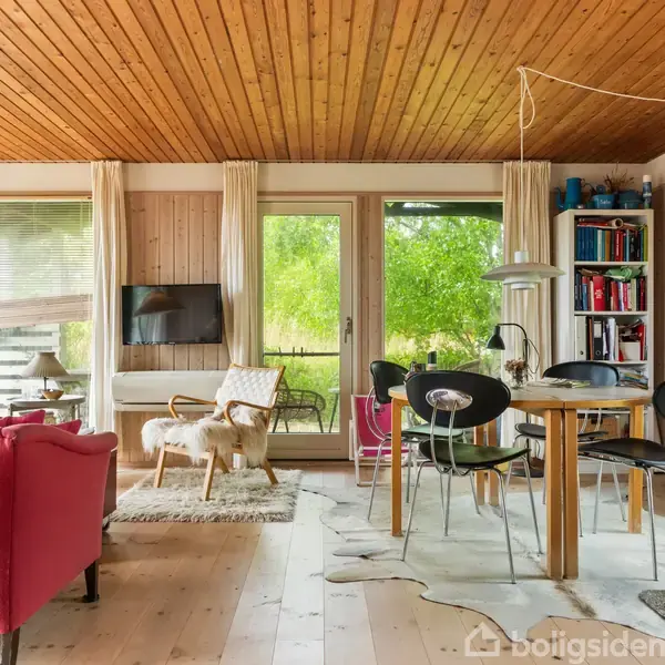 Stue med rødt sofamøbel og sort spisebordssæt, omgivet af trævægge og -loft. Et vindue og glasdør viser udsigt til en grøn natur, mens bøger fylder en reol.