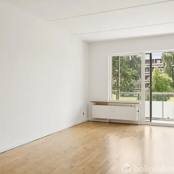 Et tomt rum med trægulv, ingen møbler, stor glasdør åbner til en balkon med udsigt til en grøn have og bygninger i baggrunden.