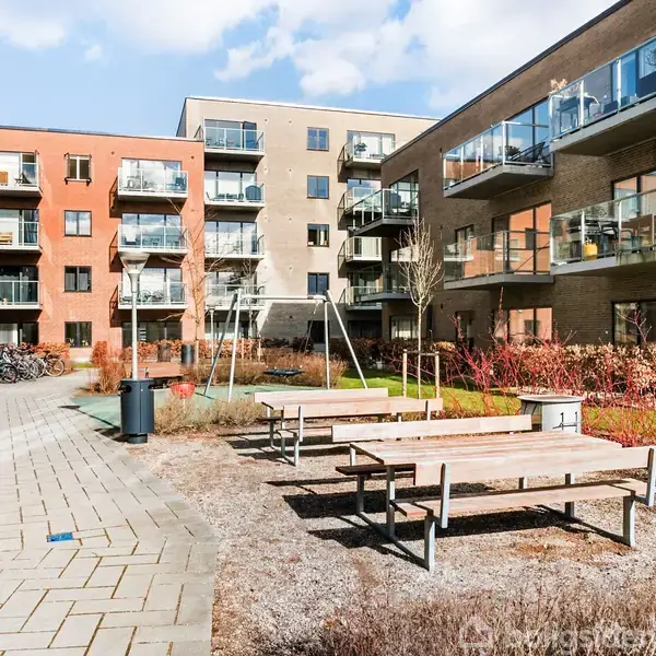 En gård med bænke og gyngestativ står stille mellem moderne lejlighedsbygninger. Den brostensbelagte sti snor sig gennem området, omgivet af planter og cykelskure.