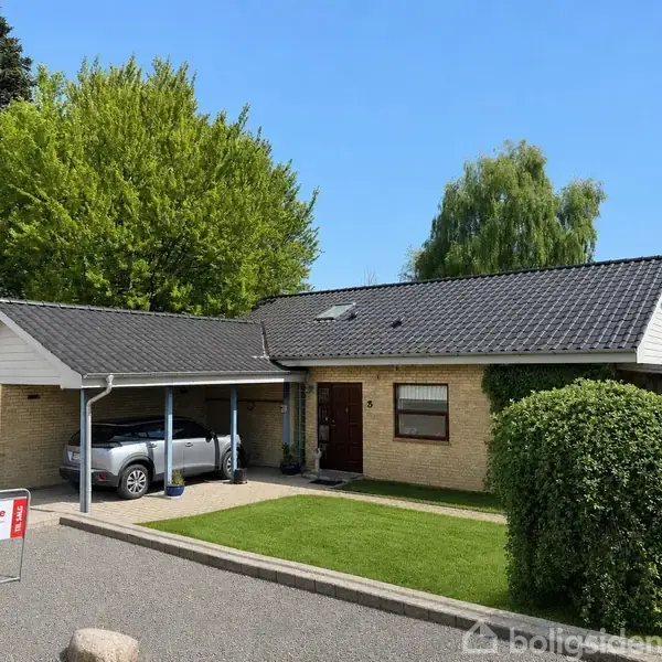 Et etplanshus af mursten med mørkt tag og indbygget carport med bil. Omkranset af grønne træer, velplejet græsplæne og indkørsel. Skilt med teksten "home" til stede.