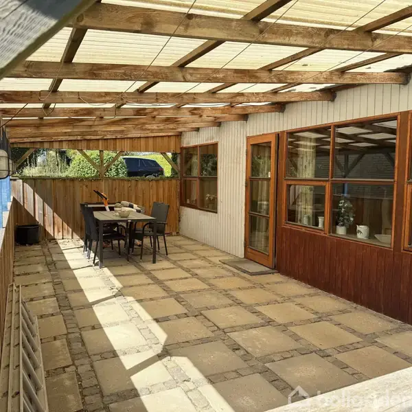 Overdækket terrasse med trærammer og glasvinduer. Et bord og stole er placeret i skyggen. Klinkebelægningsgulv giver en rustik atmosfære. Sollys filtrerer gennem det gennemsigtige tag.