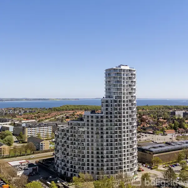 En høj, rund bygning dominerer byens skyline mod en klar blå himmel. Den omkranses af forskellige mindre bygninger og en udsigt over vandet i det fjerne.