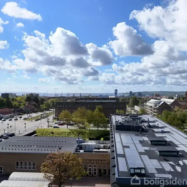 Bygning med fladt tag med solceller står stille ved siden af træer og veje, i en by med spredte bygninger under en delvis skyet himmel. Vi kan se havet i baggrunden.