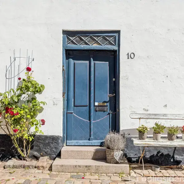 En blå dør med nummeret "10" omgivet af hvid mur, kantet af blomstrende planter og krukker på en bænk. Døren har et diamantmønster øverst, placeret ved en brostensbelægning.