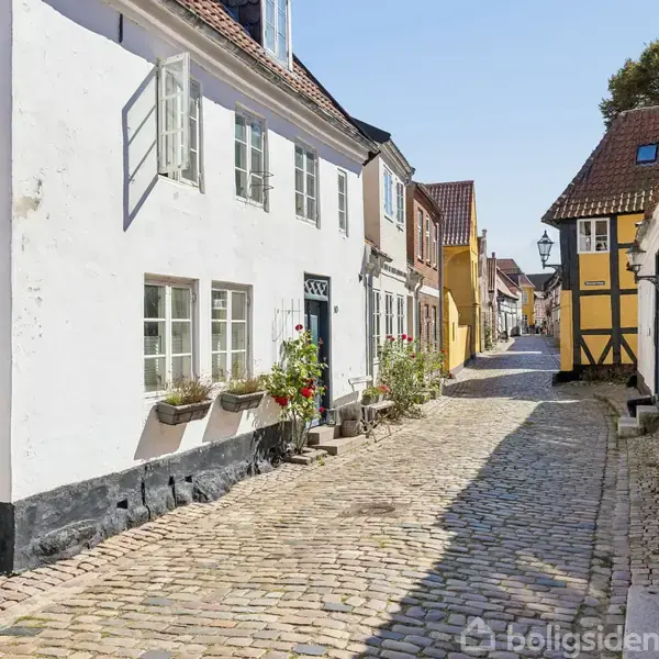 En brostensbelagt gade omgivet af charmerende gamle bygninger; en hvid facade med blomsterkasser og et gul bindingsværkshus. Solrig dag i en malerisk bymidte.