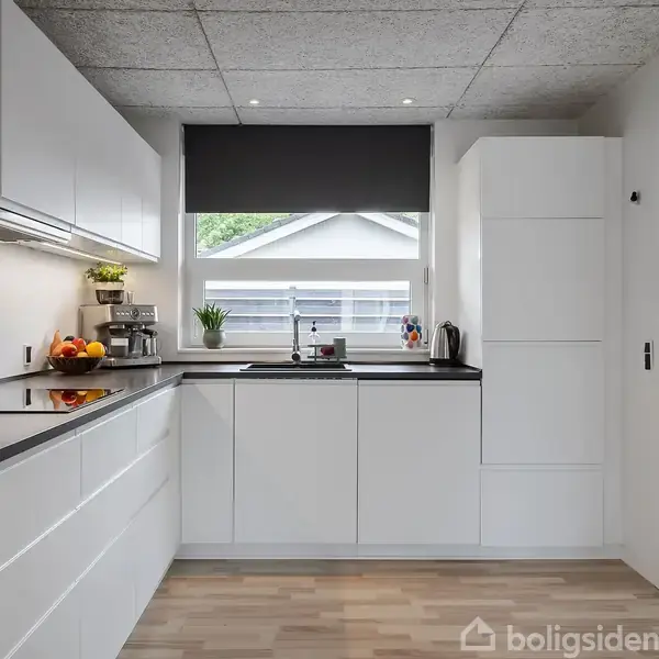 Et lyst, moderne køkken med hvide skabe og sort bordplade. En indbygget ovn og køkkenredskaber står ved en vindue med udsigt til et hus. Linoleumsgulv supplerer stilen.