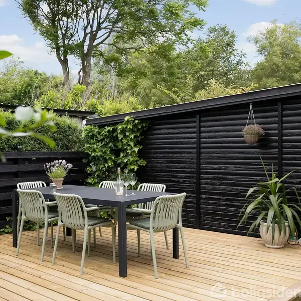 Terrassebord med fire stole står på trædæk. Bagved ses et sort træskur med planter og udsigt til grønne træer og buske. Atmosfæren er rolig og hyggelig.